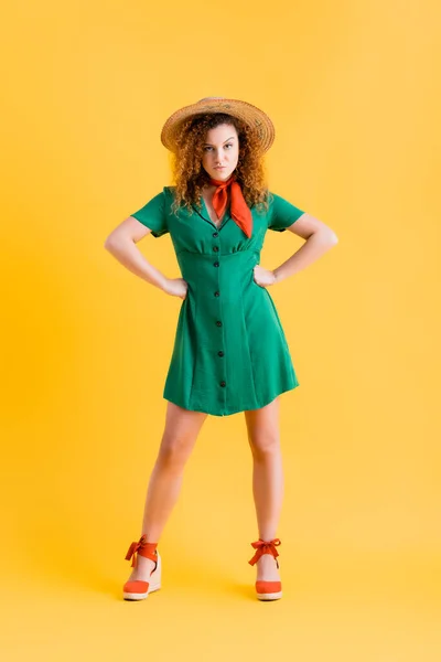 Comprimento total da mulher descontente em chapéu de palha e vestido verde de pé com as mãos em quadris em amarelo — Fotografia de Stock