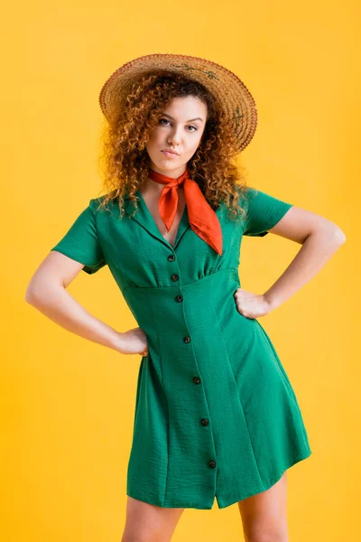 Dissatisfied woman in straw hat and green dress standing with hands on hips on yellow — Stock Photo