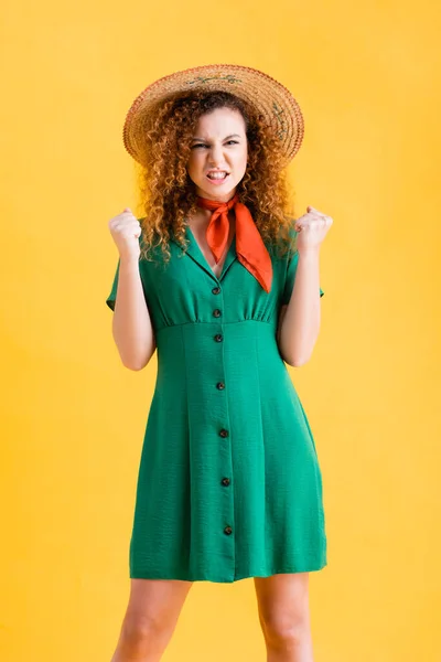 Femme en colère en chapeau de paille et robe verte debout avec les poings serrés sur jaune — Photo de stock