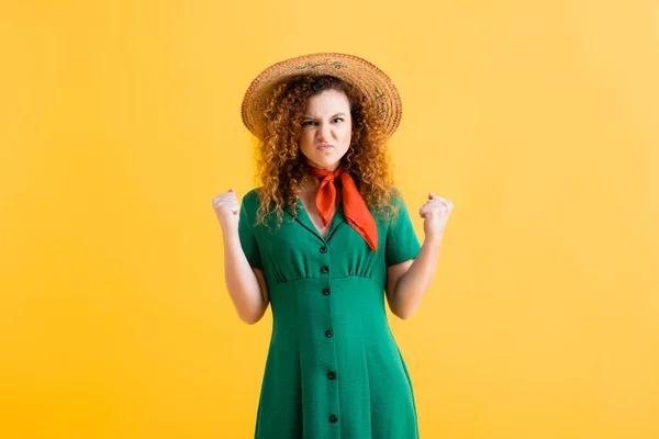 Femme irritée en chapeau de paille et robe verte debout avec des poings serrés sur jaune — Photo de stock