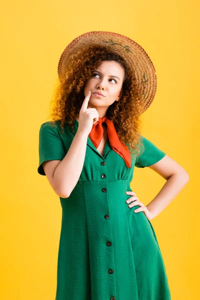 Pensive young woman in straw hat and green dress standing with hand on hip on yellow — Stock Photo
