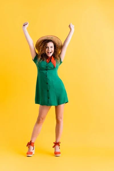 Full length of amazed woman in green dress and straw hat standing with hands above head on yellow — Stock Photo