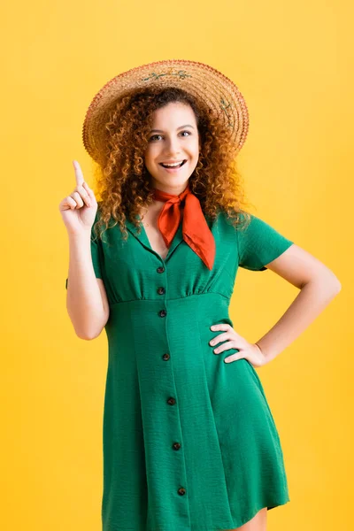 Happy young woman in straw hat and green dress pointing with finger on yellow — Stock Photo