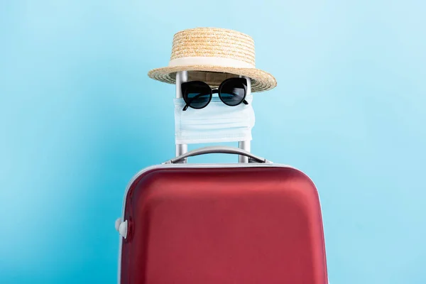 Gafas de sol, máscara médica y sombrero de paja en el equipaje rojo en azul - foto de stock