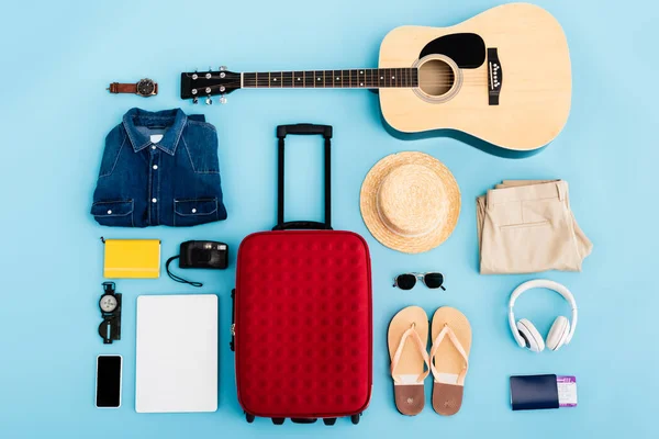 Vista superior de gafas de sol, ropa, accesorios y dispositivos cerca de equipaje rojo en azul - foto de stock