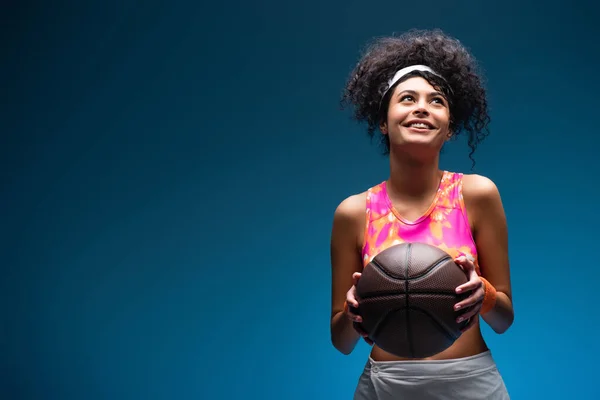 Cheerful woman in sportswear holding basketball on blue — Stock Photo