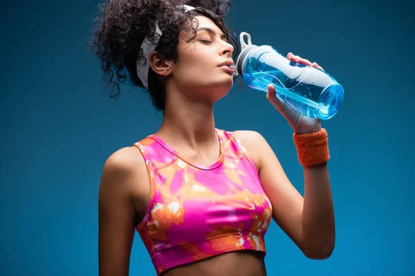 Кудрявая женщина в спортивной одежде пьет воду, держа спортивную бутылку на синем — стоковое фото