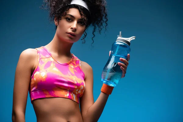 Curly woman in sportswear holding sports bottle with water on blue — Stock Photo