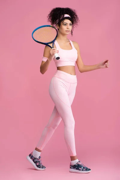 Full length of young curly sportswoman holding tennis racket while walking on pink — Stock Photo