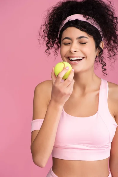 Радостная молодая женщина с закрытыми глазами, держа теннисный мяч изолирован на розовый — стоковое фото
