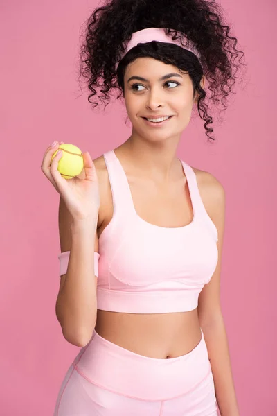 Gioiosa giovane donna guardando lontano e tenendo la palla da tennis isolata sul rosa — Foto stock