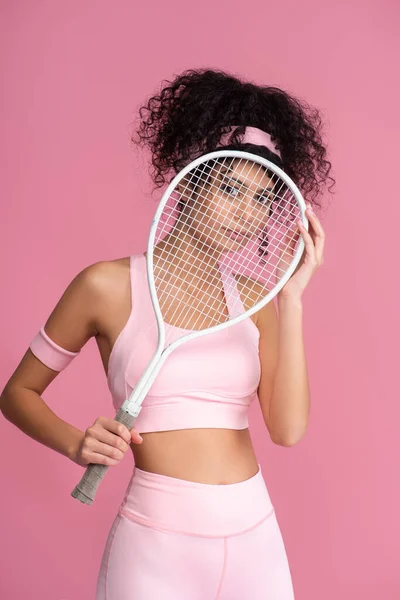 Young sportive woman looking at camera through tennis racket isolated on pink — Stock Photo