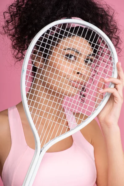 Sportive woman looking at camera through tennis racket isolated on pink — Stock Photo