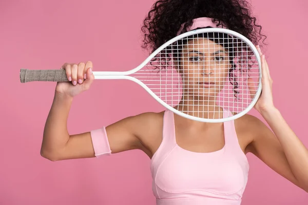 Sportive young woman looking at camera through tennis racket isolated on pink — Stock Photo