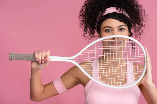 Fröhliche und sportliche Frau blickt in die Kamera, während sie den Tennisschläger isoliert auf rosa hält — Stockfoto