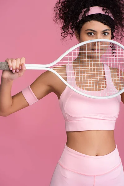 Mulher esportiva olhando para a câmera enquanto segurando raquete de tênis isolado em rosa — Fotografia de Stock