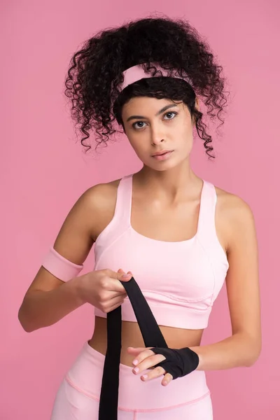 Bouclé jeune femme en vêtements de sport enveloppant la main avec la servitude isolé sur rose — Photo de stock