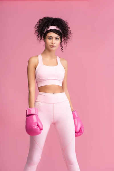 Encaracolado jovem mulher em roupas esportivas e luvas de boxe de pé isolado em rosa — Fotografia de Stock