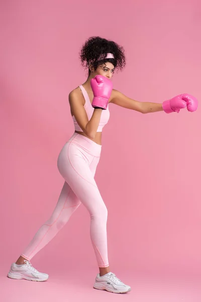 Pleine longueur de jeune femme bouclée en vêtements de sport et gants de boxe travaillant sur rose — Photo de stock