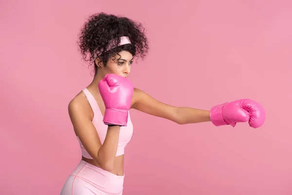 Jeune femme bouclée en vêtements de sport et gants de boxe faisant de l'exercice isolé sur rose — Photo de stock