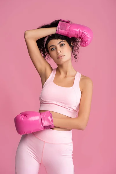 Lockige Frau in Sportbekleidung und Boxhandschuhen schaut vereinzelt in die Kamera auf rosa — Stockfoto