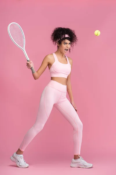 Full length of curly woman in sportswear holding racket while playing tennis on pink — Stock Photo