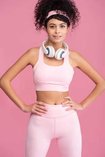 Jeune femme bouclée dans des écouteurs sans fil debout avec les mains sur les hanches isolées sur rose — Photo de stock