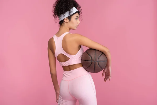 Mujer joven rizada sosteniendo baloncesto y mirando hacia otro lado aislado en rosa - foto de stock