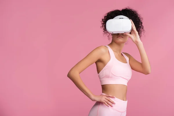 Donna riccia in realtà virtuale auricolare in piedi con mano sul fianco isolato su rosa — Foto stock
