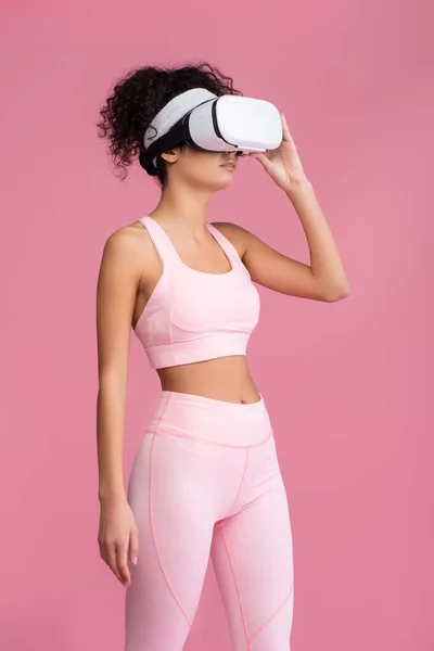Curly woman touching virtual reality headset isolated on pink — Stock Photo