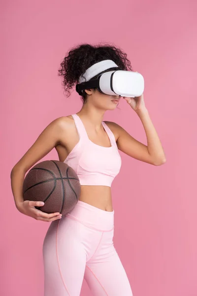 Mulher encaracolado em realidade virtual headset segurando basquete isolado em rosa — Fotografia de Stock