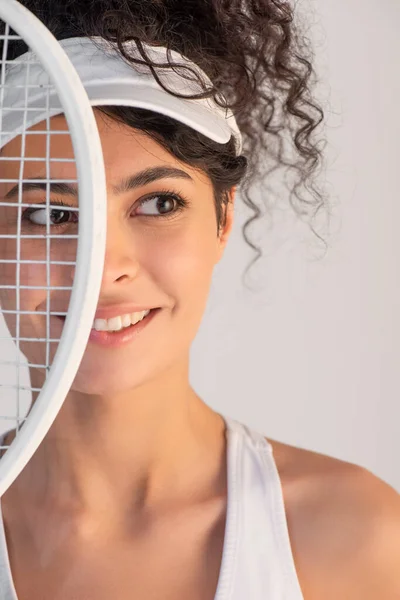 Alegre jugador en gorra mirando hacia otro lado cerca de raqueta de tenis aislado en blanco - foto de stock