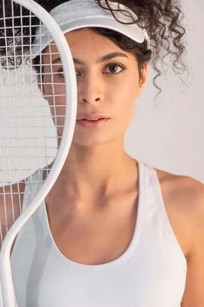 Curly sportswoman in cap looking at camera through tennis racket isolated on white — Stock Photo