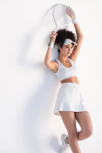 Кучерява спортсменка в кепці дивиться геть, спираючись на стіну і тримає тенісну ракетку над головою на білому — стокове фото