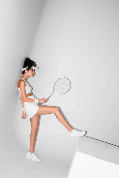 Volle Länge der lockigen Sportlerin mit Schirmmütze, die sich an die Wand lehnt, Tennisschläger hält und Würfel mit Bein auf weiß schiebt — Stockfoto