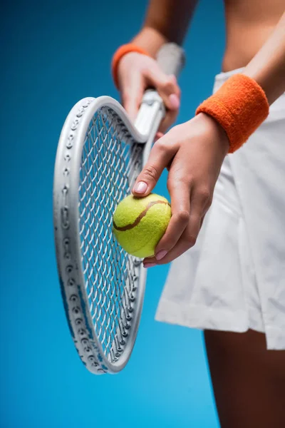 Обрізаний вид спорту молода жінка тримає тенісну ракетку і м'яч, граючи на синьому — стокове фото