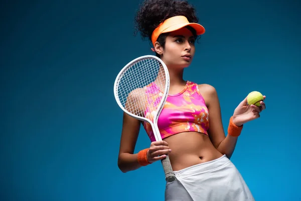 Femme sportive et bouclée tenant raquette de tennis et balle sur bleu — Photo de stock