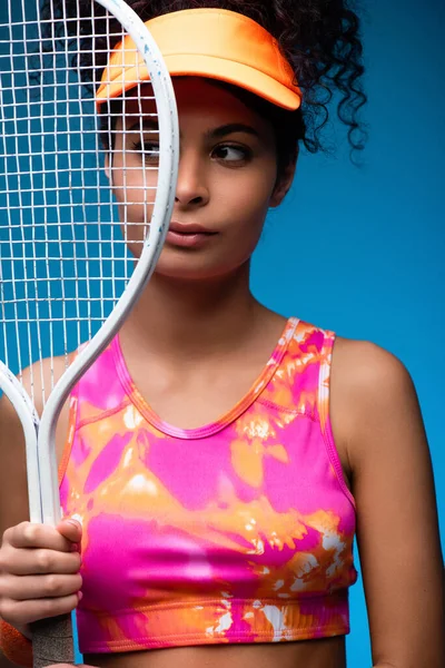 Sportive jeune femme regardant loin tout en tenant raquette de tennis sur bleu — Photo de stock