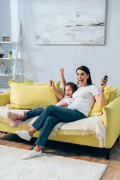 Longitud completa de madre excitada con controlador remoto e hija mirando hacia otro lado en el sofá en casa - foto de stock