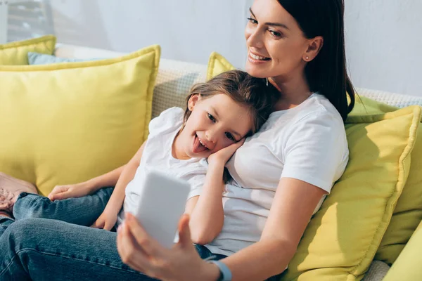 Happy mother and daughter with sticking out tongue taking selfie on couch on blurred foreground — Stock Photo