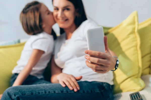 Daughter kissing smiling mother during selfie at home on blurred background — Stock Photo