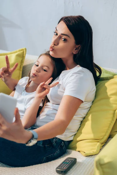 Mother with pouting lips and daughter showing peace gesture taking selfie on couch on blurred foreground — Stock Photo