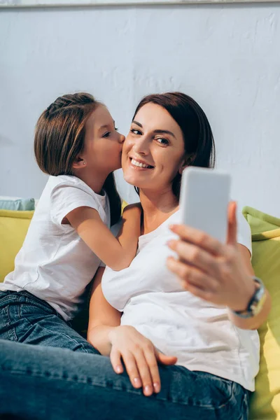 Daughter kissing mother while taking selfie on blurred foreground — Stock Photo