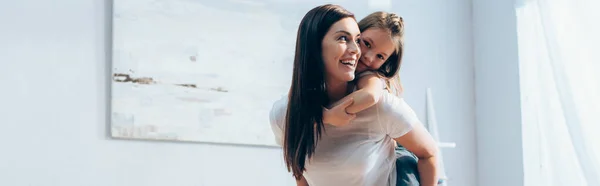 Glückliche Mutter schaut weg, während Tochter huckepack zu Hause auf verschwommenem Hintergrund, Banner — Stockfoto