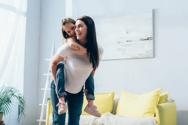 Happy mother looking away while piggybacking daughter in living room on blurred background, banner — Stock Photo