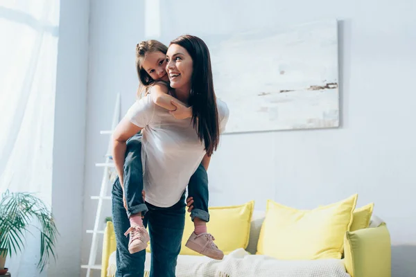 Mãe feliz olhando embora enquanto piggybacking filha na sala de estar no fundo borrado, banner — Fotografia de Stock