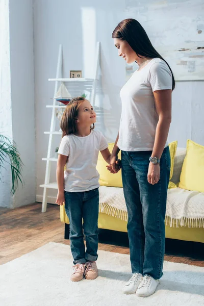 Comprimento total de sorrir mãe e filha olhando um para o outro, segurando as mãos em casa em fundo borrado — Fotografia de Stock
