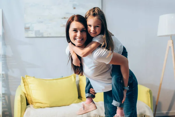 Fröhliche Mutter blickt in die Kamera, während Tochter huckepack im Wohnzimmer auf verschwommenem Hintergrund steht, Banner — Stockfoto