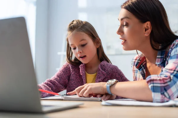 Mãe apontando com o dedo olhando para notebook perto filha apontando com lápis com laptop borrado em primeiro plano — Fotografia de Stock