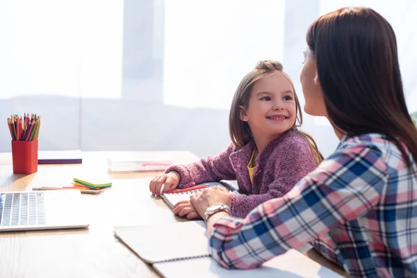 Fille joyeuse regardant la mère au bureau avec papeterie et ordinateur portable sur fond flou — Photo de stock